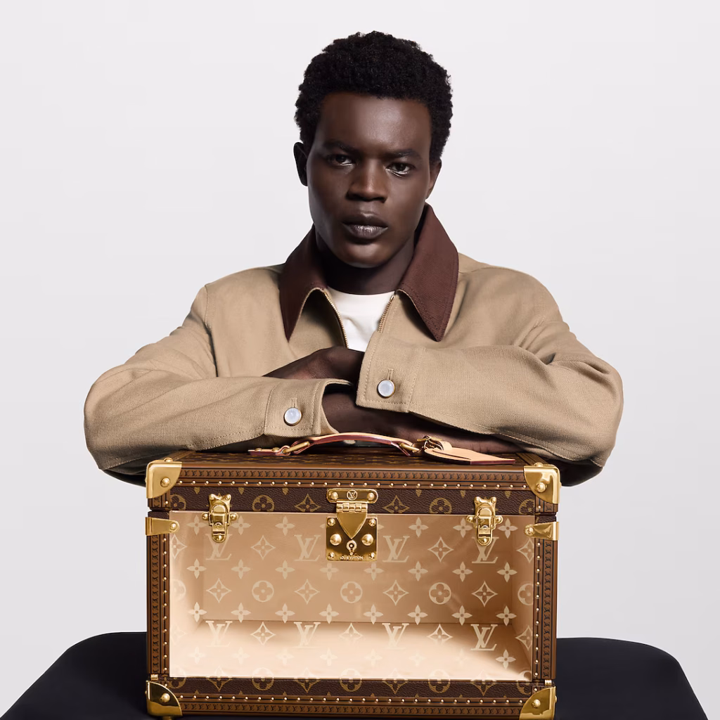 Seated model in a tan jacket resting his arms on a Louis Vuitton Shoe Box Monogram trunk with a transparent front panel.